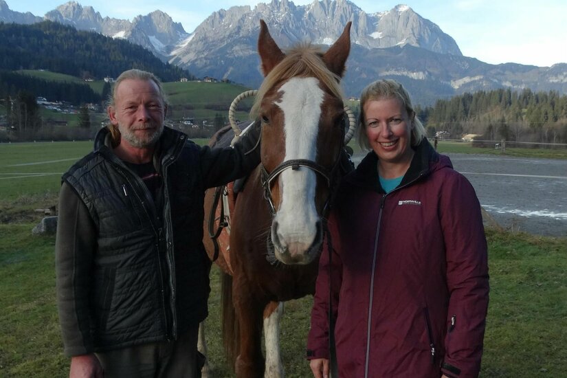 Gut Kohlhofen Reitangebot Ein Mann und eine Frau stehen neben einem braunen Pferd vor einer Bergkulisse.