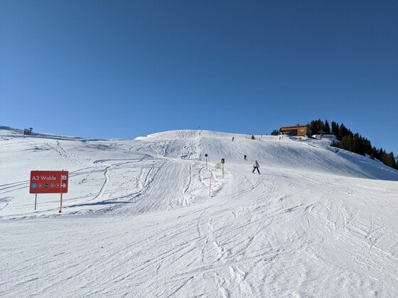 Präparierte Skipiste mit Hinweisschild und Skifahrern unter blauem Himmel.