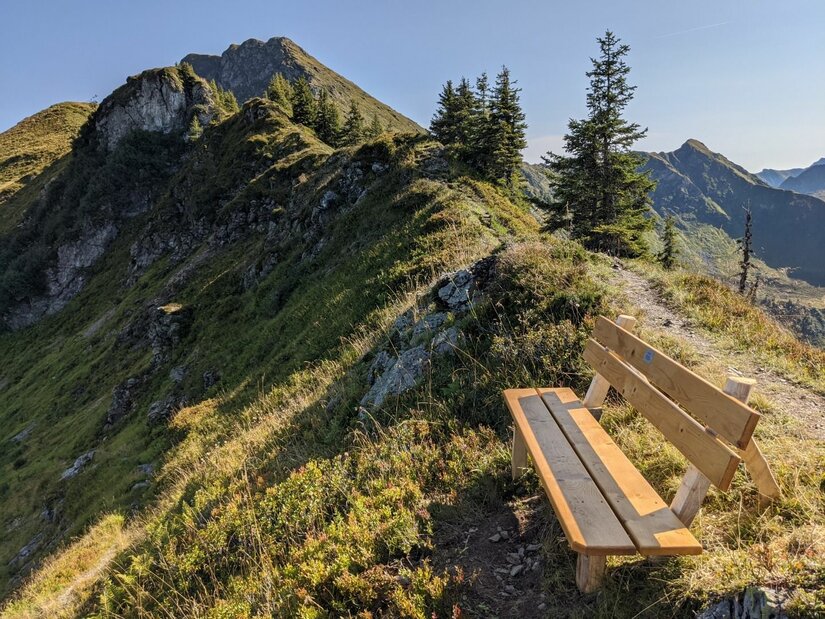 Rastbank am Berggrat Holzbank an einem schmalen Wanderpfad auf einem grasbewachsenen Bergrücken mit Blick auf Gipfel.
