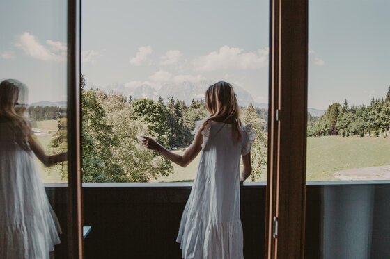 Frau in weißem Kleid blickt von einem Balkon auf eine Wald- und Berglandschaft.