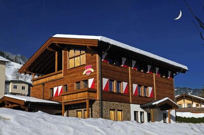 Dreistöckiges Holzhaus mit rot-weißen Fensterläden im Schnee unter blauem Himmel mit Mondsichel.