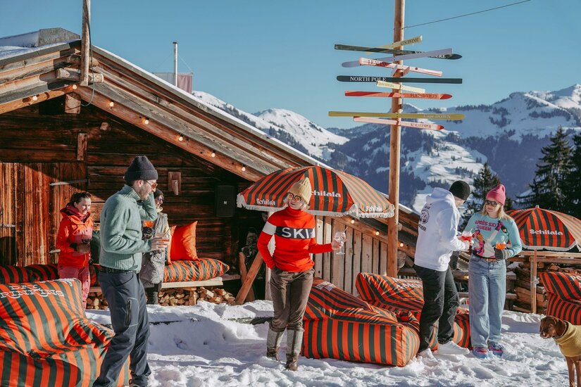 Après-Ski bei der Schilala Personen vor einer Holzhütte im Schnee mit Sitzsäcken und einem Wegweiser aus Skiern.