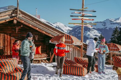 Personen vor einer Holzhütte im Schnee mit Sitzsäcken und einem Wegweiser aus Skiern.