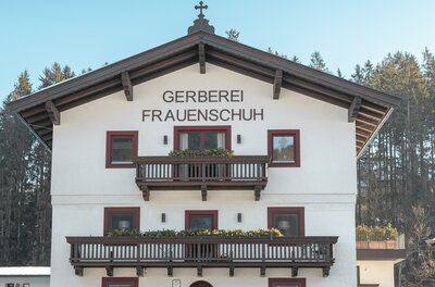 Ein traditionelles, weißes Gebäude mit einem großen Balkon und Holzverzierungen. Es trägt den Namen "Gerberei Frauenschuh" und befindet sich in einer ländlichen Umgebung.