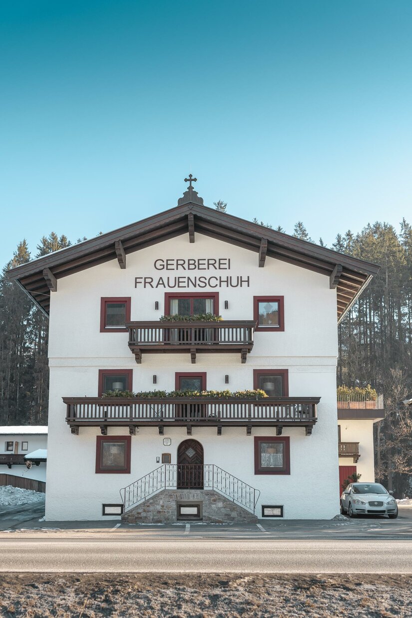 WEB_HansFrauenschuh_-66 Ein traditionelles, weißes Gebäude mit einem großen Balkon und Holzverzierungen. Es trägt den Namen "Gerberei Frauenschuh" und befindet sich in einer ländlichen Umgebung.