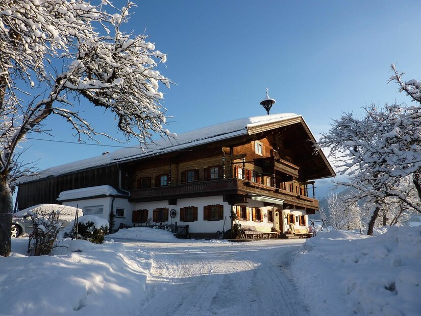 Verschneiter Bauernhof aus Holz und Stein unter blauem Himmel mit schneebedeckten Bäumen im Vordergrund.