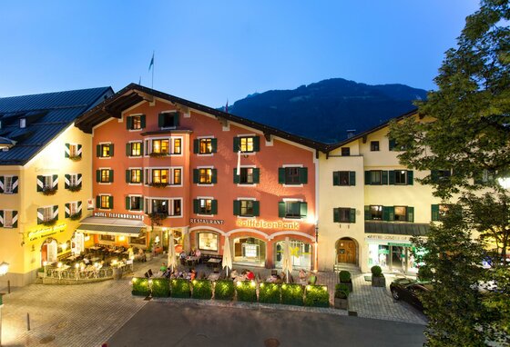 Beleuchtete Häuserfassaden am Abend in einer Stadt mit Restaurant-Terrassen und Bergen im Hintergrund.