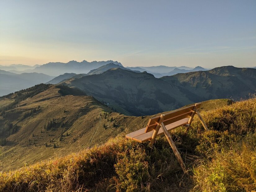 Rastplatz am Gebra Holzbank auf einem grasbewachsenen Berggipfel mit Blick auf eine weite Gebirgskette bei Sonnenuntergang.