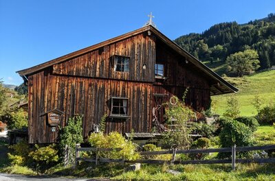Museumsweg Jochberg Altes Holzhaus mit dunkler Fassade und kleinem Gipfelkreuz vor bewaldetem Hang.
