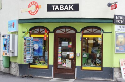Außenansicht einer Tabak-Trafik mit hellgrüner Fassade, Postkartenständer und Verkaufsautomaten.