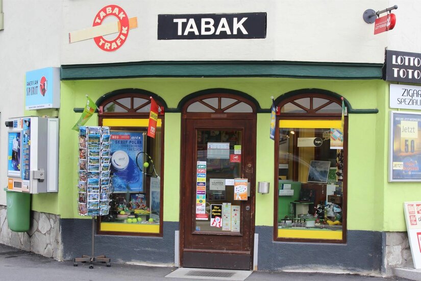 Außenansicht einer Tabak-Trafik mit hellgrüner Fassade, Postkartenständer und Verkaufsautomaten.