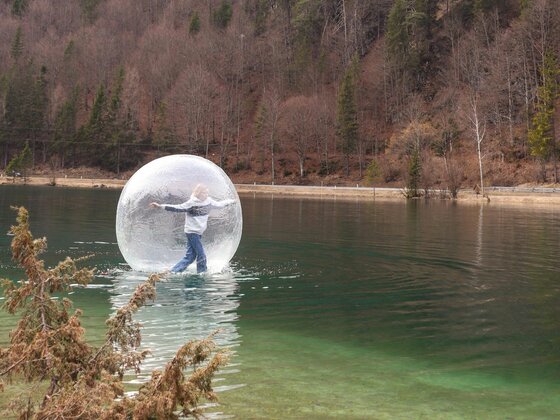 Eine Person steht in einer transparenten Ballon-Kugel auf einem ruhigen See. Im Hintergrund sind Bäume und sanfte Hügel zu sehen.