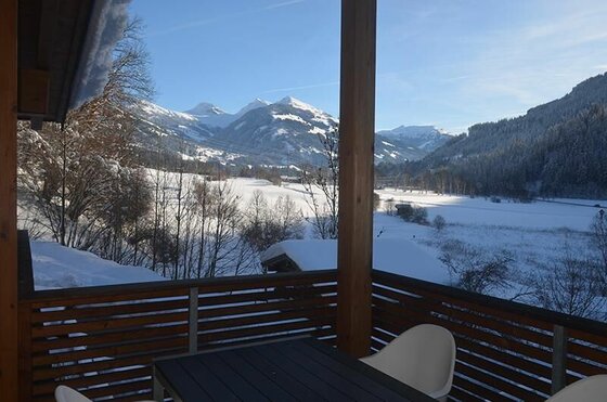 Blick von einem Holz-Balkon auf ein verschneites Tal und schneebedeckte Berggipfel unter blauem Himmel.