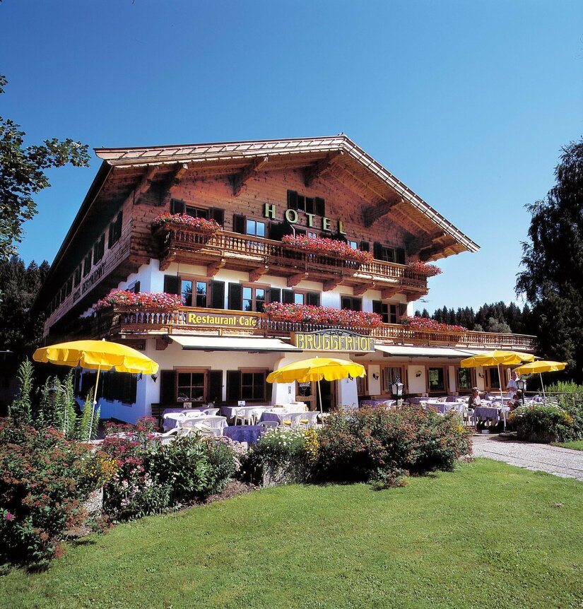 Traditionelles Holzhotel mit Restaurantterrasse, gelben Sonnenschirmen und Blumenschmuck unter blauem Himmel.