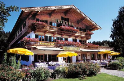Traditionelles Holzhotel mit Restaurantterrasse, gelben Sonnenschirmen und Blumenschmuck unter blauem Himmel.
