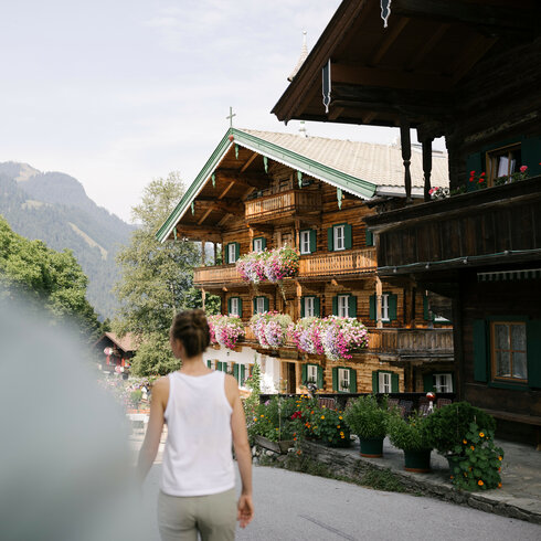 Das Bild fängt eine Frau ein, die auf einem Weg zu einem einladenden Gasthaus mit üppigen Blumenarrangements in den Alpen geht.