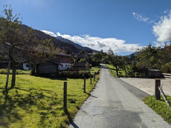 Asphaltierter Weg führt an Holzhütten und grünen Wiesen in Jochberg vorbei.