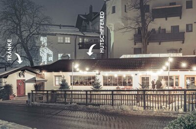 Ein gemütliches Restaurant bei Nacht, beleuchtet von warmen Lichtern. Im Hintergrund sind einige Häuser und Bäume zu sehen.