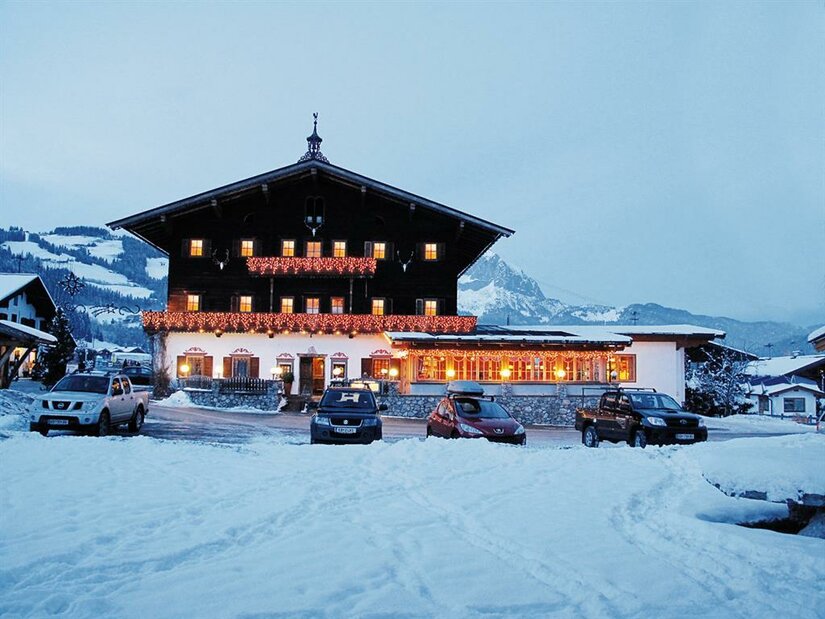 Ein beleuchteter Landgasthof in Holzbauweise bei Dämmerung in einer verschneiten Berglandschaft mit parkenden Autos.