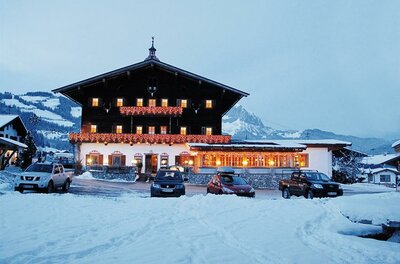Ein beleuchteter Landgasthof in Holzbauweise bei Dämmerung in einer verschneiten Berglandschaft mit parkenden Autos.