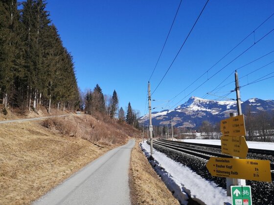 Radweg an der Bahnstrecke Asphaltierter Weg neben Bahngleisen mit gelben Wegweisern und schneebedeckten Bergen im Hintergrund.