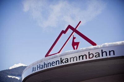 Hahnenkammbahn Ein Schild der Hahnenkammbahn mit einem stilisierten Dach und einem Reh. Der Schreibstil ist modern und ansprechend, vor einer bergigen Kulisse.