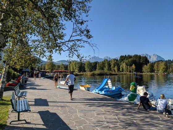 Uferpromenade am Schwarzsee Uferpromenade an einem See mit Tretbooten, Parkbänken und Spaziergängern vor bewaldetem Hintergrund.