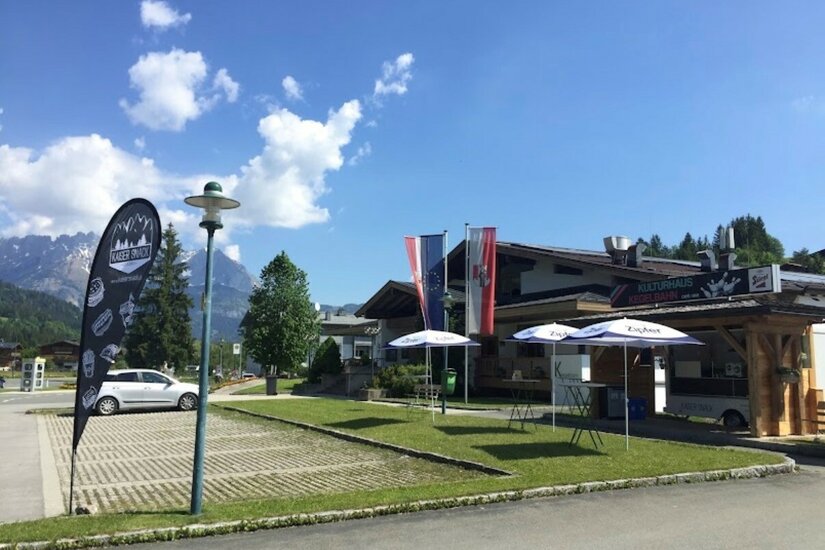 Kulturhaus und Kaisersnack Außenansicht des Kulturhauses mit Imbissstand, Sonnenschirmen und Parkplatz vor Bergpanorama.