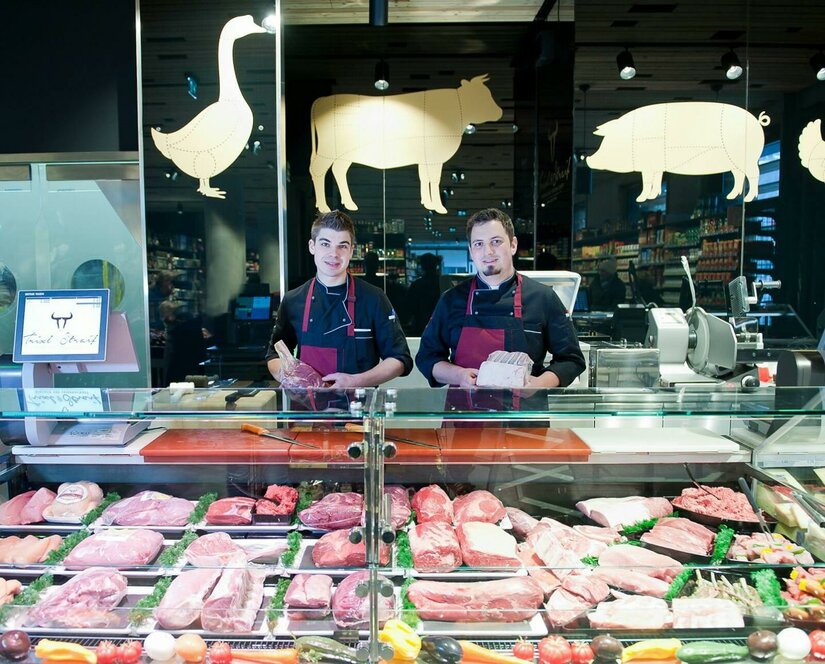 Metzgerei Trixl und Straif Zwei Metzger hinter einer Verkaufstheke mit verschiedenen Fleischsorten.