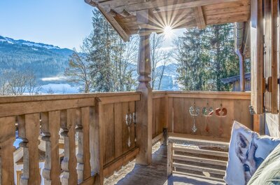Holzbalkon eines Chalets mit Blick auf verschneite Berge und Wälder bei Sonnenschein.