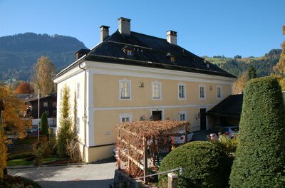 Ein historisches Haus mit gelber Fassade und einem schönen Garten. Im Hintergrund sind Berge und ein blauer Himmel sichtbar.