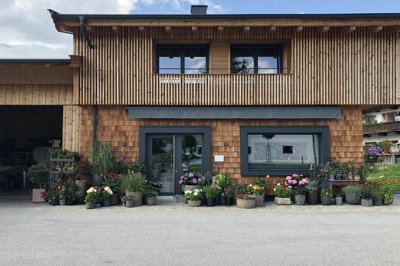 Modernes Holzgebäude mit Schindelfassade und zahlreichen Blumenkübeln vor dem Eingang.