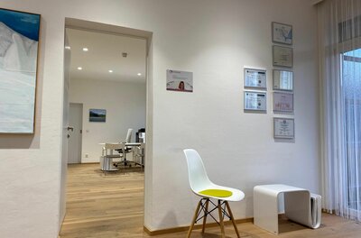 Wartebereich der Praxis Heller Warteraum mit weißem Stuhl, Parkettboden und Blick in ein angrenzendes Büro.