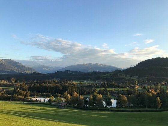 Alpines Talpanorama Blick über grüne Wiesen und Wälder auf ein Bergpanorama unter blauem Himmel mit leichten Wolken.