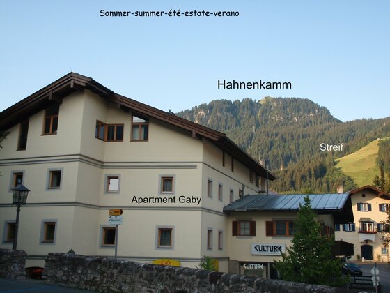 Hausansicht Apartment Gaby Mehrstöckiges helles Gebäude mit Satteldach vor bewaldetem Berghang unter blauem Himmel.