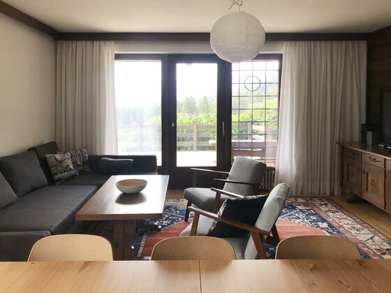 Wohnzimmer Hahnenkamm im Haus Astrid Wohnzimmer mit grauem Ecksofa, Holztisch, zwei Sesseln und großem Fenster zur Terrasse.