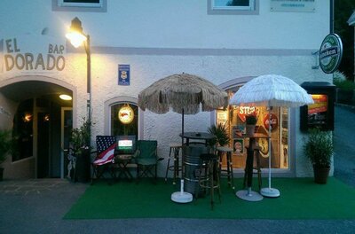 Außenbereich der Bar El Dorado mit Bastschirmen, Stehtischen und Barhockern vor einer weißen Hauswand.