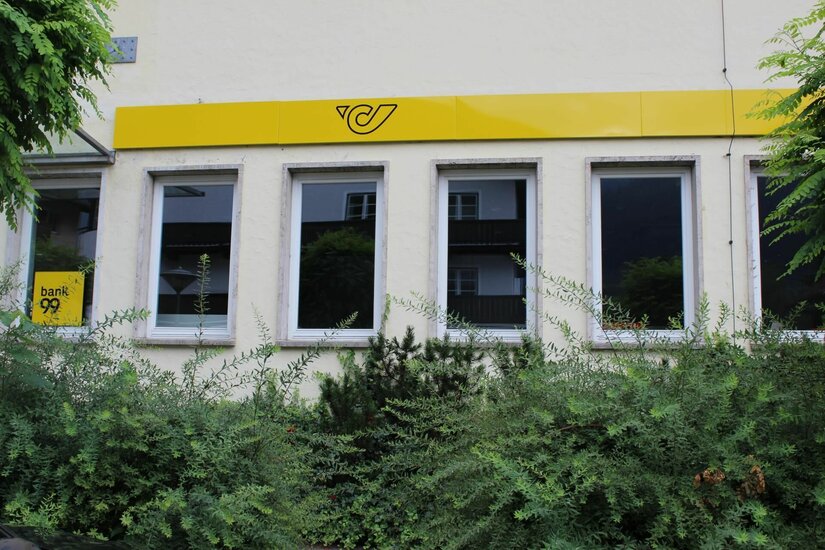 Außenansicht eines Postamtes mit gelbem Logo über den Fenstern und grünen Sträuchern im Vordergrund.