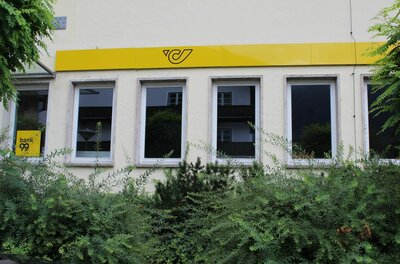 Postamt Kitzbühel Außenansicht eines Postamtes mit gelbem Logo über den Fenstern und grünen Sträuchern im Vordergrund.