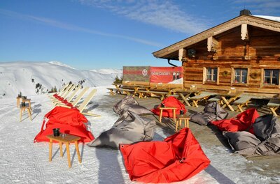 Sonnenterrasse der Bärenbadalm mit roten und grauen Sitzsäcken im Schnee vor einer Holzhütte.