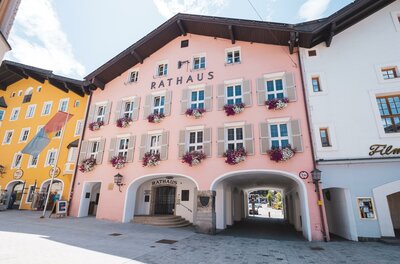 Ein farbenfrohes Rathaus in einer malerischen Straße. Die Fassade ist rosa mit Blumenfensterbänken und umgeben von weiteren schönen Gebäuden.