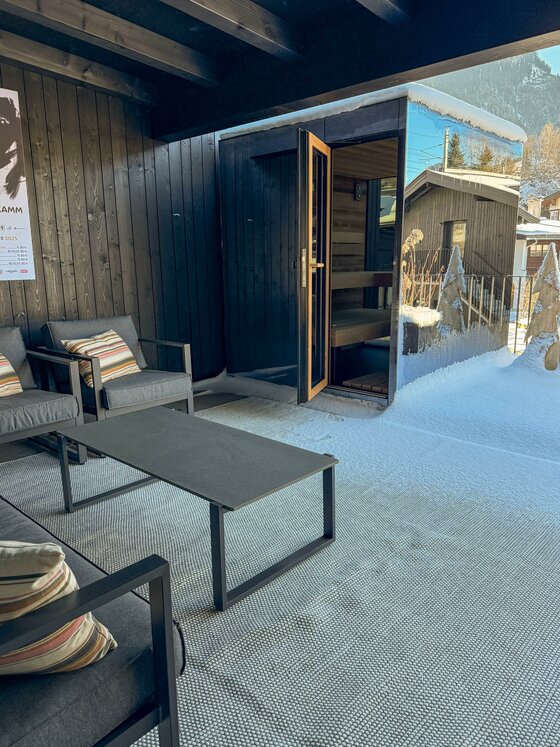 Eine gemütliche Terrasse mit modernen Möbeln und Blick auf eine verschneite Umgebung. Im Hintergrund ist eine Sauna zu sehen, die einladend geöffnet ist.