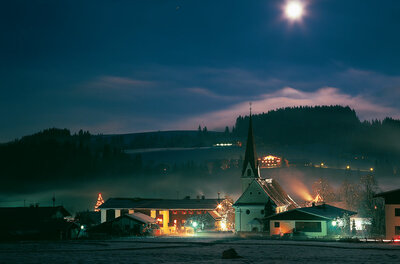 Beleuchtetes Dorf mit Kirche bei Nacht unter Vollmond und leichtem Nebel.