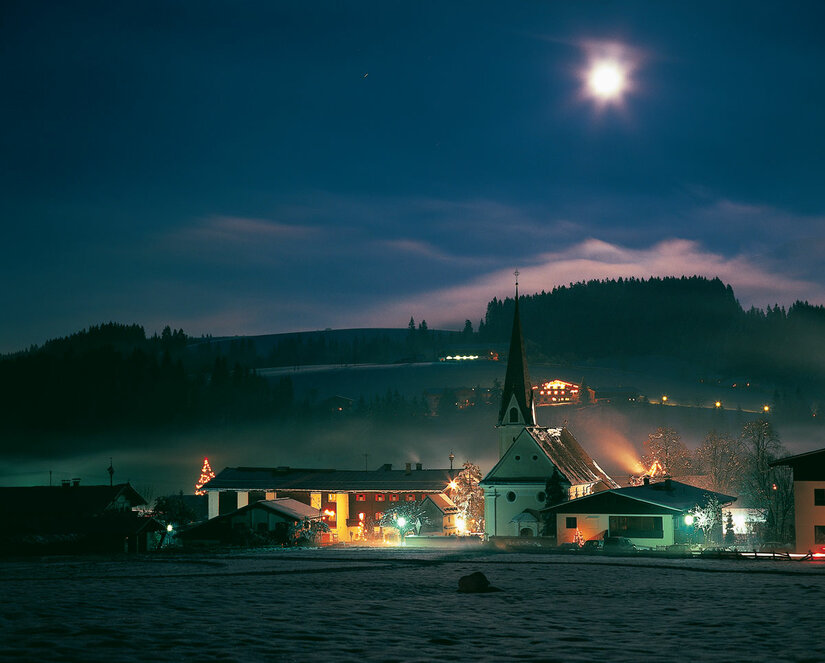 Beleuchtetes Dorf mit Kirche bei Nacht unter Vollmond und leichtem Nebel.