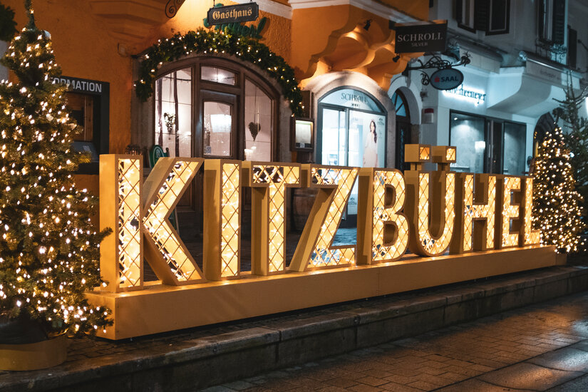 Ein großer, leuchtender Schriftzug 'Kitzbühel' steht in einer festlichen Straße, umgeben von Weihnachtsdekoration und Lichtern.