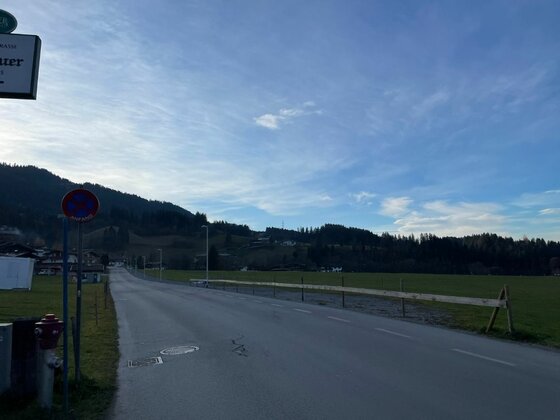 Radweg Wilder Kaiser Asphaltierte Straße führt an einer grünen Wiese vorbei in Richtung bewaldeter Berge unter blauem Himmel.