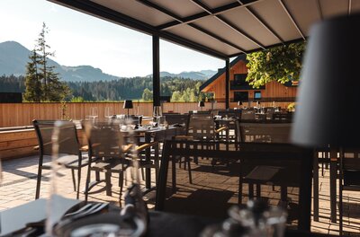 Überdachte Restaurantterrasse mit gedeckten Tischen, Blick auf Nadelwälder und Berge.