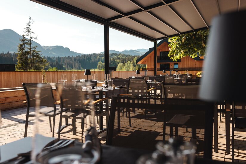 Überdachte Restaurantterrasse mit gedeckten Tischen, Blick auf Nadelwälder und Berge.