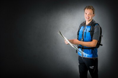 Reini Percht Ein junger Mann steht mit einem Rucksack und hält eine Landkarte in der Hand. Er lächelt und trägt eine blaue Weste.
