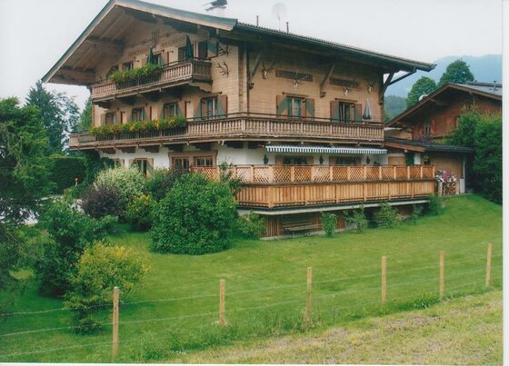 Mehrstöckiges Holzhaus im alpinen Stil mit Balkonen, Blumenkästen und einer großen Terrasse vor einer Wiese.